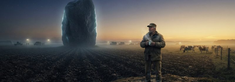 A vast, dark plowed field in rural Germany at the edge of dawn, with a massive monolithic object resting silently on the earth — dark as wet slate, roughly the size of a church, faintly glowing with a slow, rhythmic pulse of pale blue-white light along its surface. A lone farmer stands in the foreground in work clothes, holding a mug of tea, watching from a distance as golden morning light begins to crawl across the furrows toward the object. A ring of distant military vehicles and floodlights is barely visible at the horizon. A small herd of cattle grazes calmly nearby. The atmosphere is deeply still, neither threatening nor triumphant — suffused with quiet awe and uncertain wonder. The sky transitions from deep indigo to warm amber at the horizon. Cinematic, photorealistic style with a slightly desaturated, cold-toned palette contrasted by warm sunrise light, evoking the mood of literary science fiction — intimate, grounded, and profoundly human.
